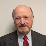 Head-and-shoulders portrait of an older man with glasses, gray blazer, white shirt, and red tie, against a neutral background.