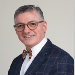 Smiling man in a dark plaid suit, white shirt, and bow tie against a light background (professional headshot)