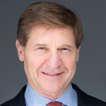Head-and-shoulders portrait of a smiling middle-aged man with light brown hair, wearing a blue shirt and dark blazer, against a gray backdrop.