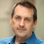 Portrait of a middle-aged man in a blue shirt looking at the camera in an офис-like setting.