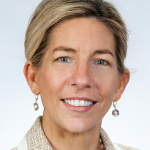 Professional headshot of a smiling woman with blonde hair, wearing a beige blazer and pearl earrings, suitable for a team page.