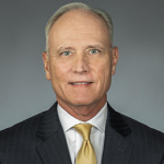 Professional headshot of a man in a dark pinstripe suit, white shirt, and gold tie, facing the camera against a gray background.