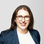 Professional headshot of a smiling woman with brown hair, glasses, wearing a navy blazer and white top.