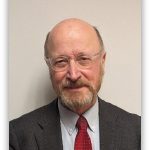 Portrait of an older man with glasses, balding head and gray beard, wearing a dark suit and red tie, looking at the camera.