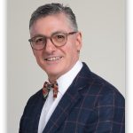 Portrait of a smiling man in a dark plaid suit, bow tie, and glasses, facing slightly to the right.