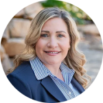 Professional headshot of a smiling woman in a navy blazer and striped blue shirt, outdoors with a stone background.