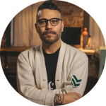 Portrait of a man with glasses and a beige cardigan, arms crossed, in a cozy office with a green logo on the sleeve.