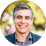 Portrait of a smiling man with gray hair wearing a blazer, outdoors with blurred greenery in the background.